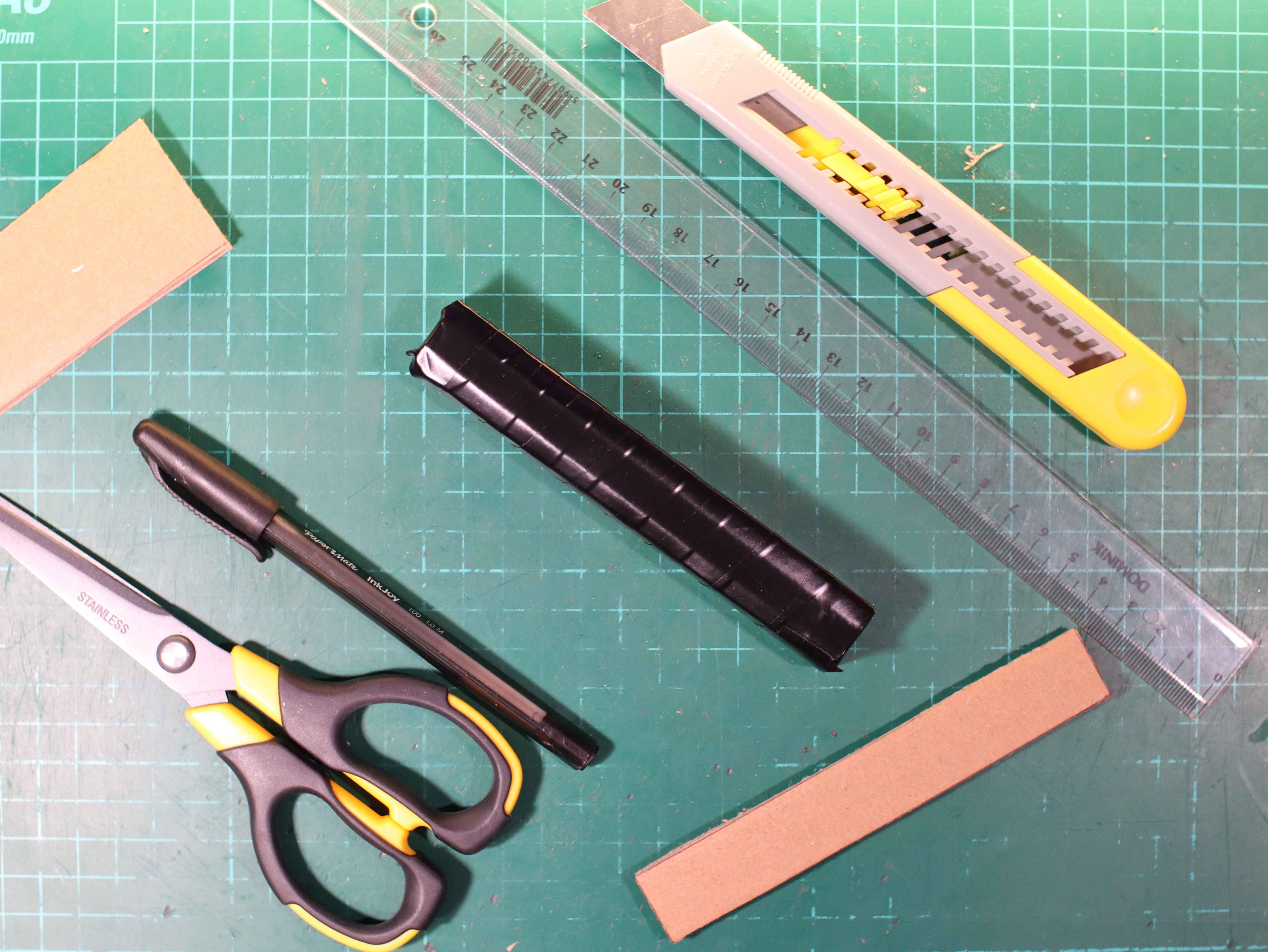 Cardboard battery covered with black insulating tape. Scissors, a knife, a pen, a ruler are on the green service mat.