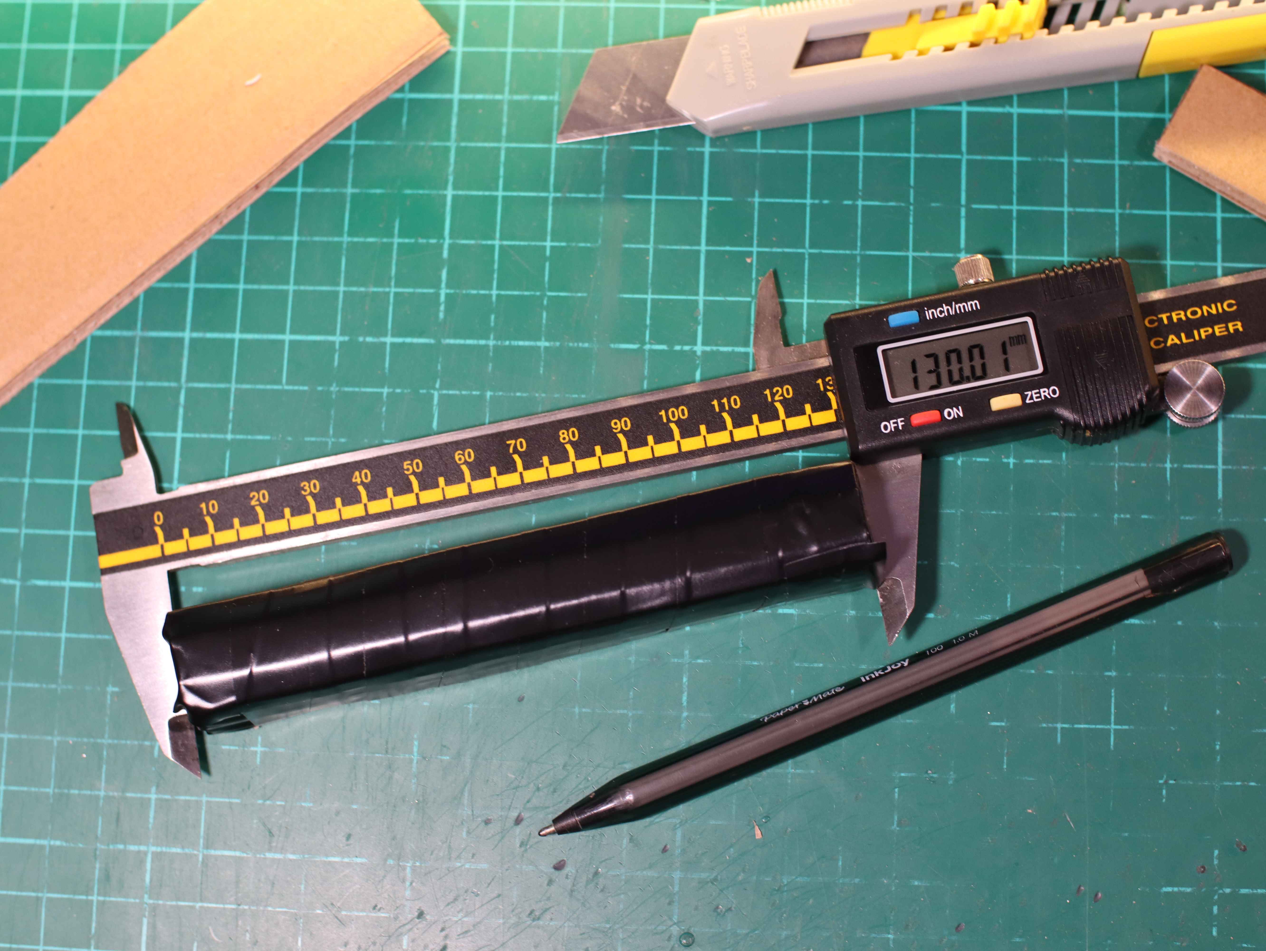 Measurement of the length of the airsoft battery model from the cardboard. The model is covered with black insulating tape, the caliper, knife and pen lie on the service mat.