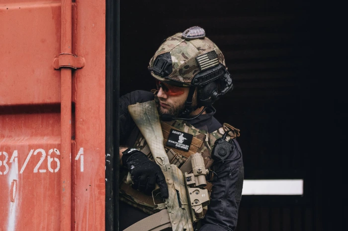 Osoba w taktycznym stroju, w hełmie moro z naszywką flagi USA, wychylająca się zza rogu dużego, czerwonego kontenera. 