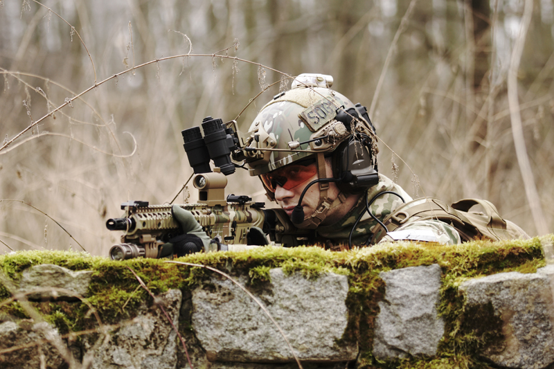 un joueur d'airsoft portant un casque et des lunettes de protection vise une réplique, cachée derrière un mur.